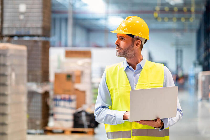 Worker wearing a hardhat and safety vest holding a laptop in a warehouse