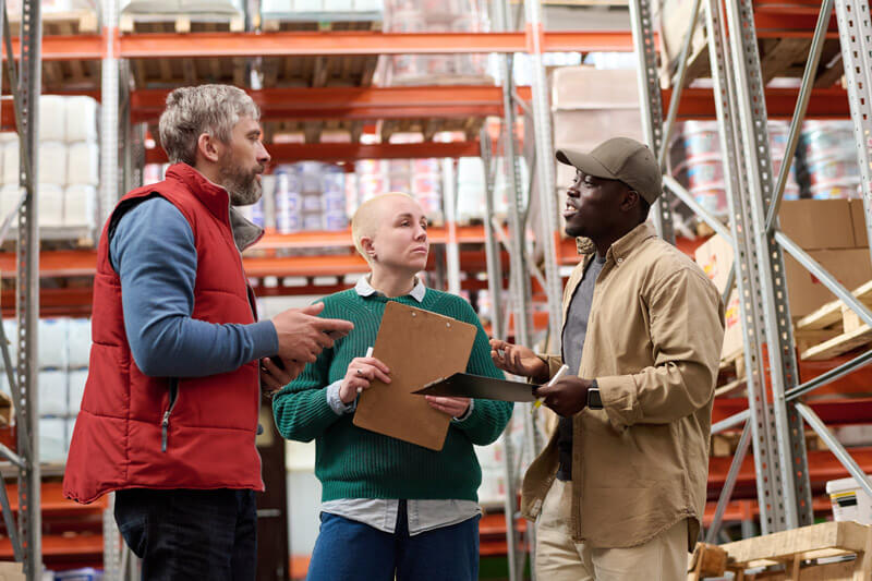 Three workers, two holding clipboards, talking in a warehouse